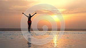 Back view, silhouette of woman in swimsuit, standing and relaxing with hands up, on edge of outdoor infinity pool with
