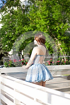 back view of a slender woman in a blue short dress with a full skirt standing at the white wooden railing of a summer