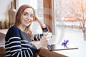 Back view smiling beautiful young woman read book with digital tablet near big window in a cafe or home. Attractive