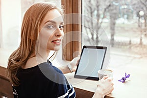 Back view smiling beautiful young woman read magazine with digital tablet near big window in a cafe or home. Attractive