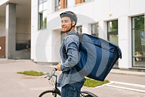 Back view of smiling handsome young delivery man in protective helmet posing standing near bicycle in city street on