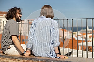 Back view of smiling tourist couple sitting bench on old city street and looking each other