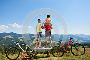 Back view of sporty family cyclists standing on wooden bench, resting after cycling bicycles