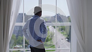 Back view of stressed African American man throwing out paperwork sighing looking out the window in home office