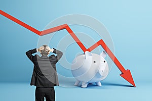 Back view of stressed businessman with falling red chart arrow with piggy bank on blue background. Economic recession,