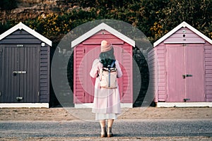 Back view stylish hipster woman with color hair in total pink outfit and backpack looking at wooden beach huts. Off