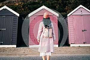 Back view stylish hipster woman with color hair in total pink outfit and backpack looking at wooden beach huts. Off photo