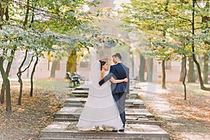 The back view of the stylish newlyweds hugging and walking in the sunny park.