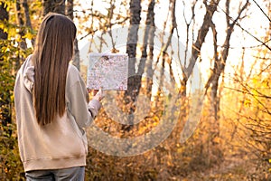 Back view of stylish woman holding paper map, wearing warm beige sweater and jeans, looking at forest view while