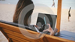 Back view successful businesswoman with drink works on laptop financial diagrams in lounge chair on amazing ocean beach.