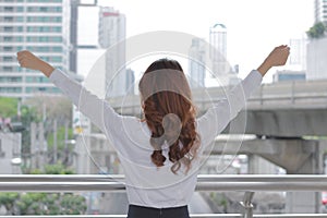 Back view of successful young Aian business woman raising her hands at urban building city background.