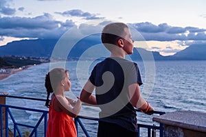 Back view of tanned preteen boy brother little girl sister standing at iron fence looking at beach sea waves in evening.