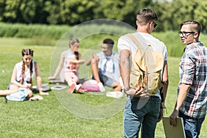 back view of teenage boys walking in park while classmates studying behind