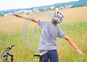 Back view of teenager boy in protective helmet spread his arms out like a bird standing next to his bike in park on