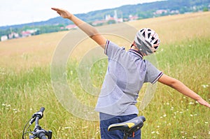 Back view of teenager boy in protective helmet spread his arms out like a bird standing next to his bike in park on