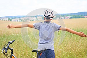 Back view of teenager boy in protective helmet spread his arms out like a bird standing next to his bike in park on