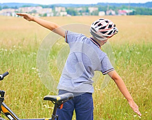 Back view of teenager boy in protective helmet spread his arms out like a bird standing next to his bike in park on