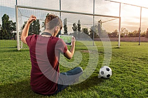 Back view, Teenager sitting on football/soccer field watching the game on tablet