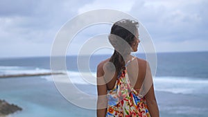 Back of view of thoughtful young brunette woman with long hair wearing long dress standing on a rock by the ocean