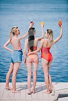 back view of three young women in swimwear holding glasses with summer cocktails