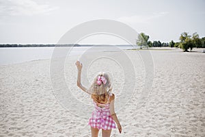 Back view to young blonde girl look like a Barbie doll in pink tones waving her hand on beach