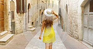 Back view of tourist woman holding hat and walking in typical medieval Tuscany street in Italy. Panoramic banner view