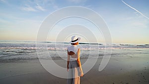 Back view of tourist woman in white summer hat standing at the beautiful beach
