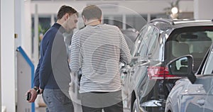 Back view of two adult Caucasian men standing next to the row of automobiles and talking. Friends choosing new car in