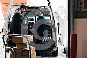 Back view of two delivery people checking information on a digital tablet while standing at van trunk near a warehouse