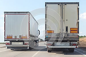 Back view of two dump trucks drives out of town on a flat asphalt road among fields against a blue sky with clouds on a sunny day