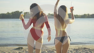 Back view of two joyful young slim women dancing with drink bottles at sunset on beach. Cheerful Caucasian tourists