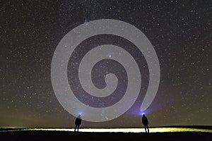 Back view of two men with head flashlights on green grassy field under dark blue summer starry sky.