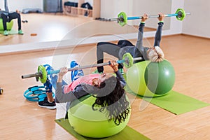 Back view of two women lifting barbell lying on stability ball while exercising in gym