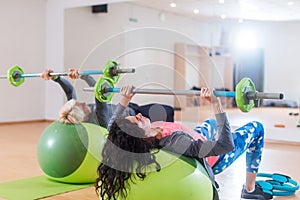 Back view of two women lifting barbell lying on stability ball while exercising in gym