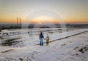 Back view of two young children in warm clothing standing in frozen snow field holding hands on copy space background of setting