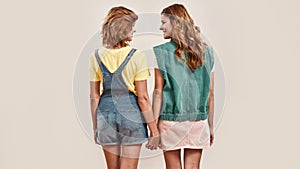 Back view of two young girls, twin sisters in casual wear holding hands, looking at each other, posing together