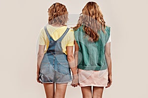 Back view of two young girls, twin sisters in casual wear holding hands, posing together, standing  over light