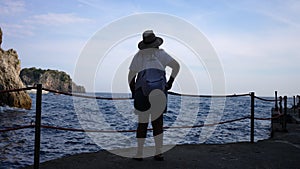 Back view of unrecognizable female tourist in hat with backpack standing enjoying view of beautiful seascape of
