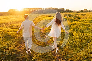 Back view of unrecognizable happy young couple in love taking each other hand and walking in green meadow towards golden