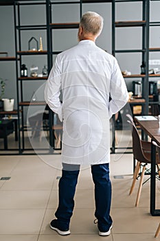 Back view of unrecognizable senior adult male doctor in white medical uniform standing in hospital office.