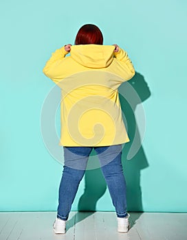 Back view of a voluptuous lady in jeans and bright yellow hoodie putting capuche on her head.