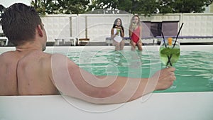 Back view of wet young man getted out from the open-air swimming pool on the background of two beautiful girls. A