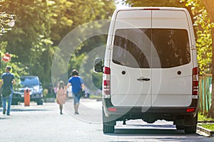 Back view of white passenger medium size commercial luxury minibus van parked n shadow of green tree on summer city street i with