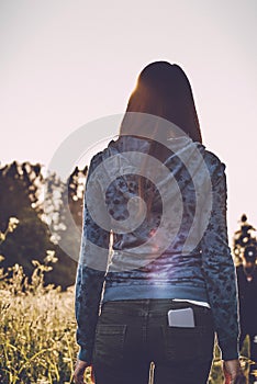 Back view of woman in casual wear enjoying the beauty of sunny autumn day in high grass. Young female in hoodie and jeans standing
