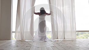 Back view of the woman covered in the bedsheet walking toward the floor-to-ceiling window and looking out from behind