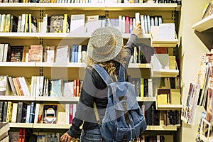 Back view of woman with hat and backpack choosing a book to buy inside a library or newspaper store. People prepare to travel.