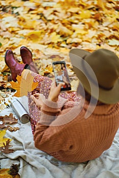 Back view of woman in hat taking photo on camera phone while relaxing in autumn park