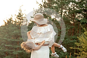 Back view of woman mother carrying little girl daughter in arms near pines in park forest illumined by sunset in summer.