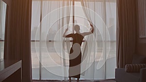 Back view of woman opening curtains and looking out of window to cityscape at hotel in the morning. Modern buildings at