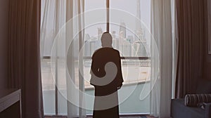 Back view of woman opening curtains and looking out of window to cityscape at hotel in the morning. Modern buildings at